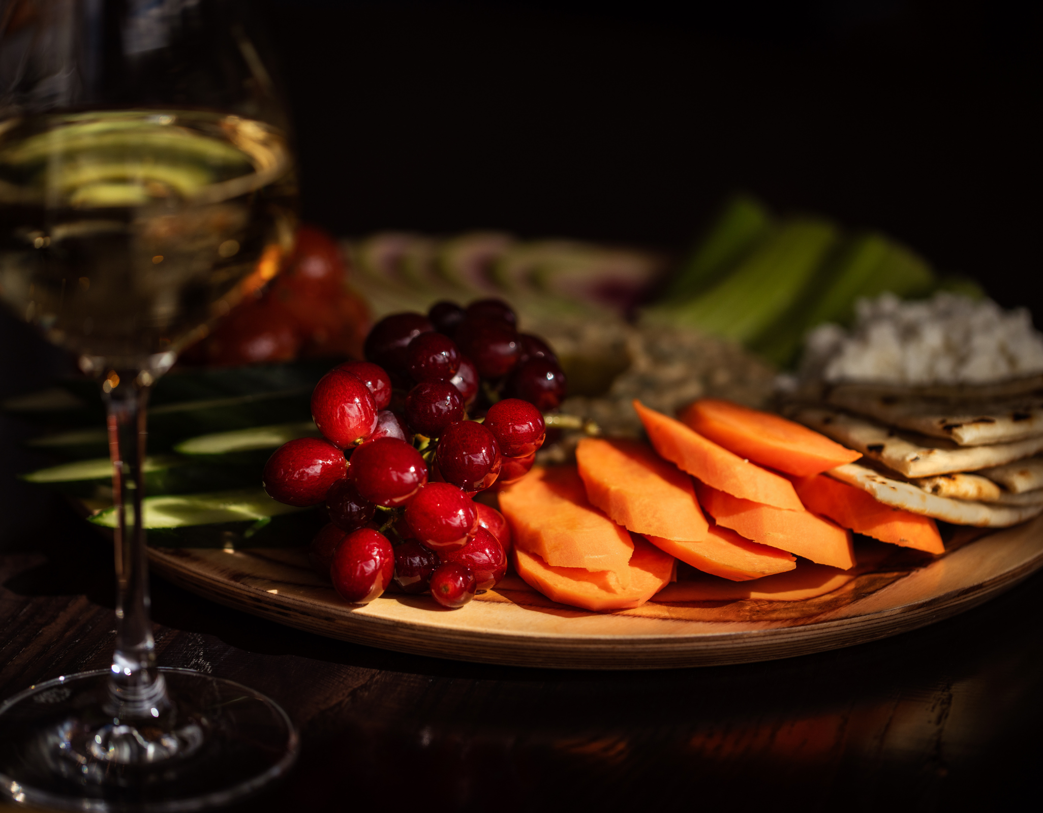 A close-up of a charcuterie board featuring red grapes, sliced melon, cucumbers, crackers, and cheese, with a glass of white wine in the foreground under warm, low lighting.