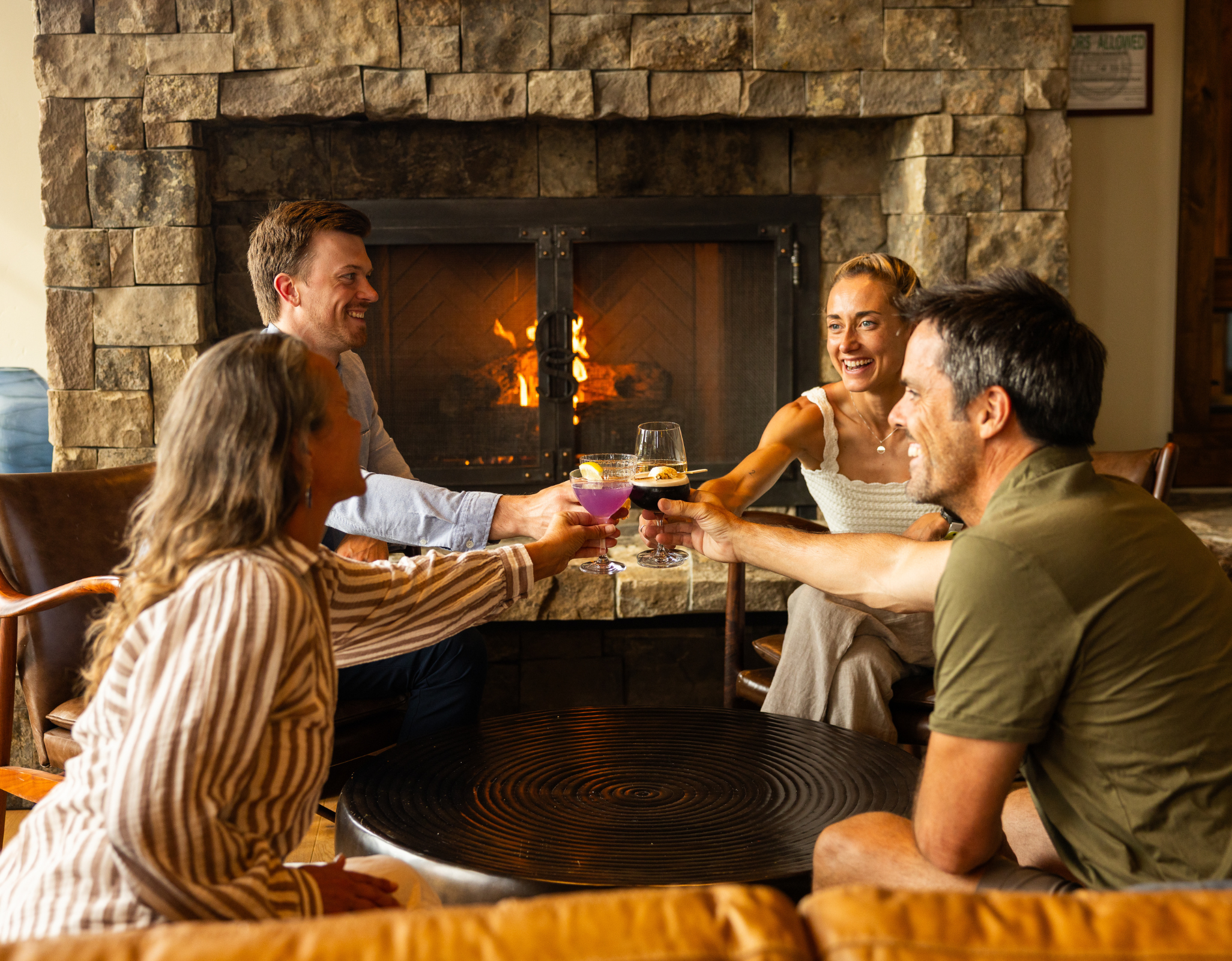 Four adults sit together around a table in front of a stone fireplace, smiling and toasting drinks in a warm, cozy indoor setting.