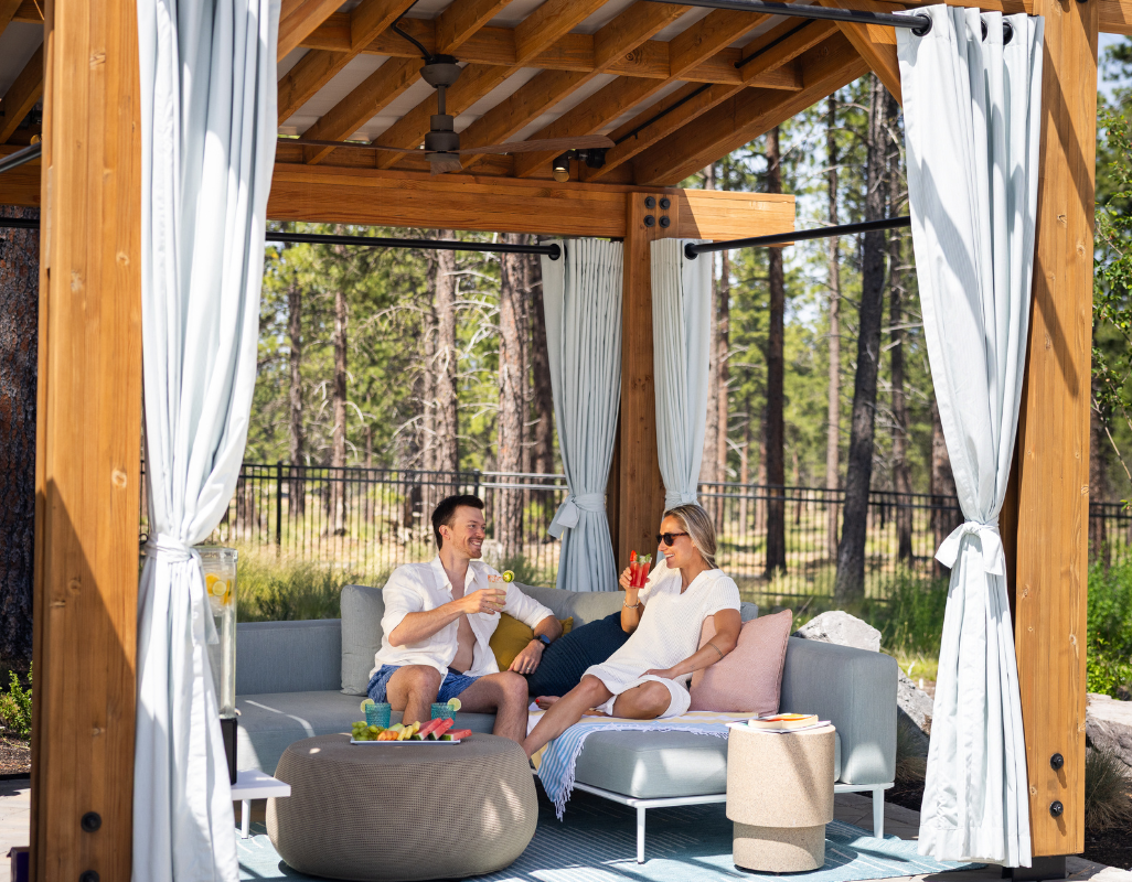 A couple relaxes in an outdoor cabana surrounded by tall pine trees, sitting on a cushioned sectional sofa and enjoying drinks. The wooden structure features soft curtains, a ceiling fan, and a cozy seating area with a small table holding fruit and refreshments.