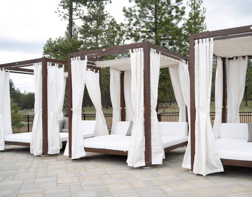 A row of outdoor cabana daybeds with wooden frames and white curtains sits on a paved patio, set against a backdrop of tall pine trees and open natural surroundings.