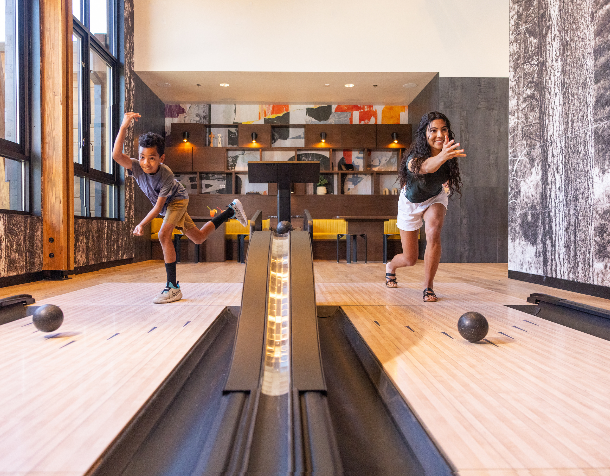 A woman and a child play duckpin bowling indoors, each mid-throw on adjacent lanes. The space features warm wood flooring, large windows letting in natural light, and a modern lounge area with seating and abstract wall art in the background.