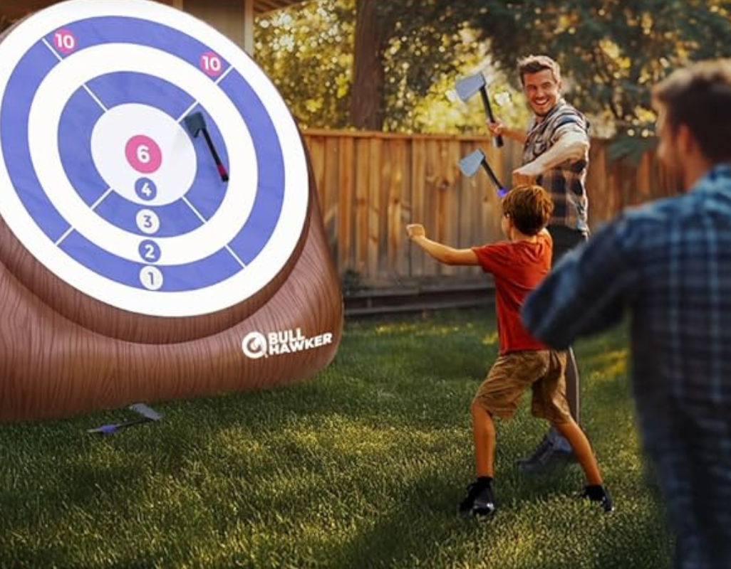 A child throws a foam axe at a backyard target while two adults watch and smile, enjoying an outdoor lawn game together.