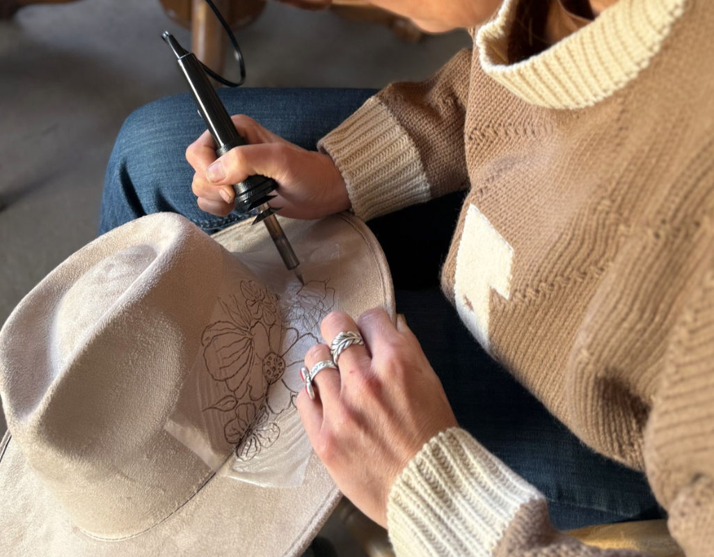 Person using a handheld tool to carefully burn a floral design into a light-colored felt hat while seated, wearing a cozy sweater and rings.