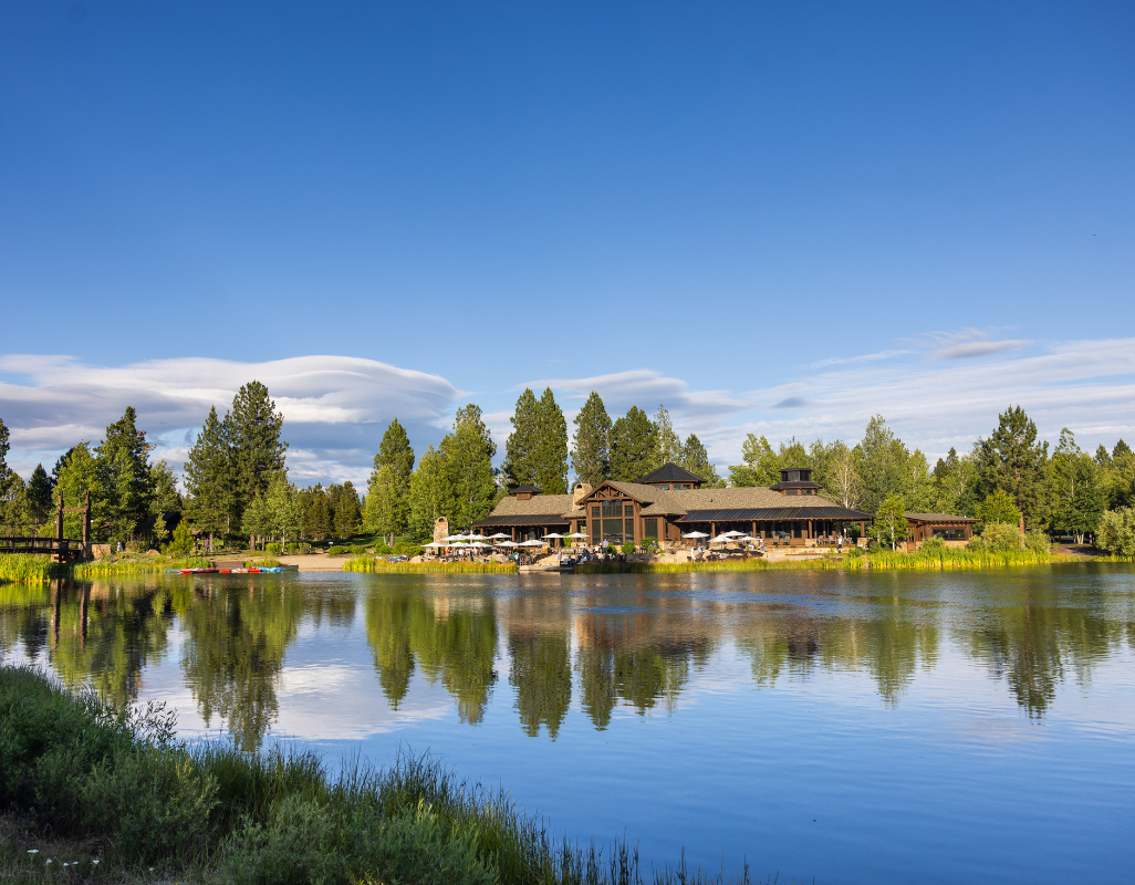 Lakeside lodge with outdoor seating and umbrellas, surrounded by trees and reflected in calm water under a clear blue sky.