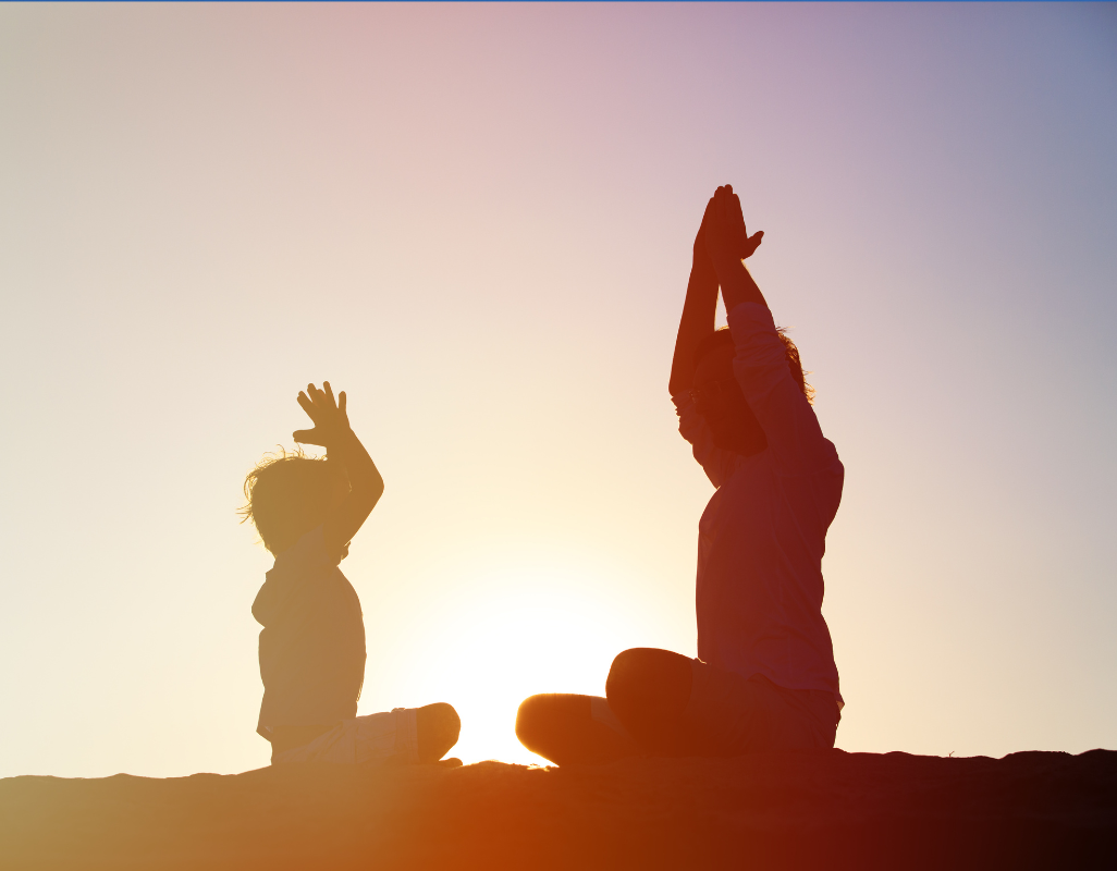 Silhouetted adult and child sitting cross-legged outdoors at sunset, raising their arms in a calm, yoga-like pose against a glowing sky.