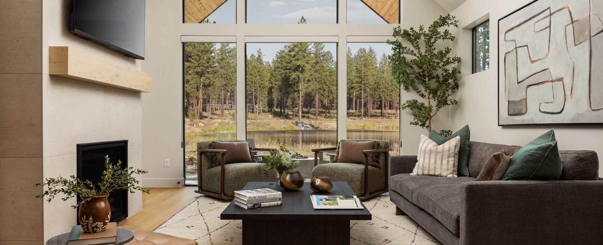 Modern living room with vaulted windows overlooking a wooded lakeside setting, featuring a fireplace, dark sofa, upholstered armchairs, and a large coffee table.