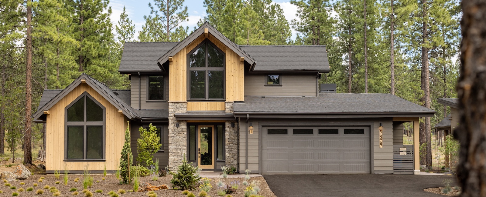 Contemporary two-story mountain home among pine trees with natural wood and gray siding, stone accents, dramatic gabled windows, and an attached two-car garage.