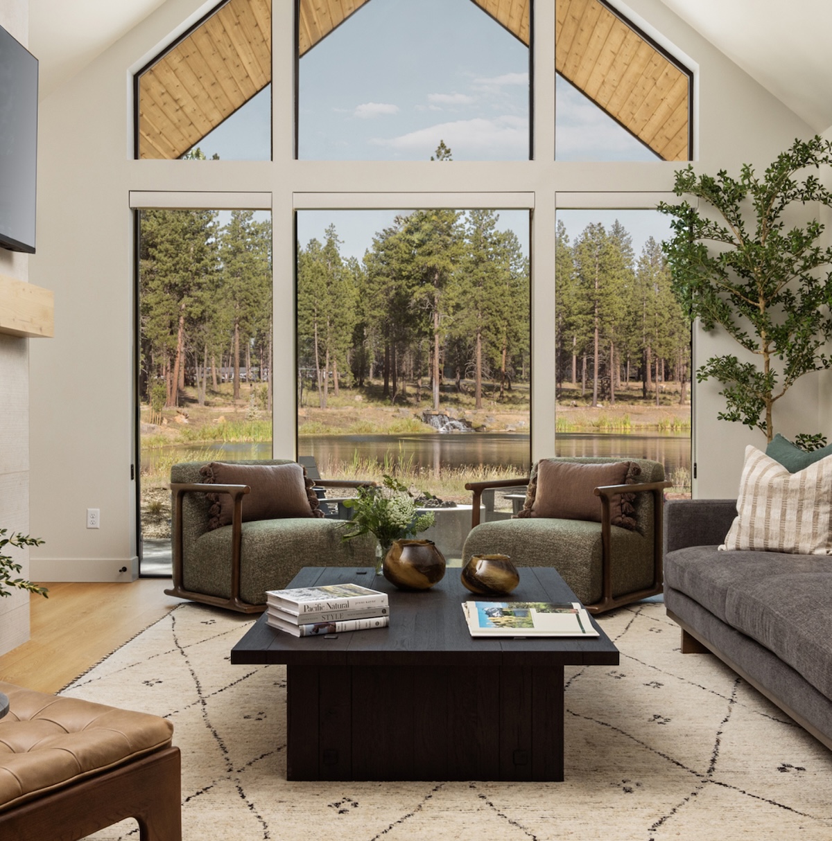 Modern lakeside living room with a gray sectional, two green armchairs, a dark wood coffee table, fireplace, large windows overlooking a pine forest and pond, and neutral contemporary decor.