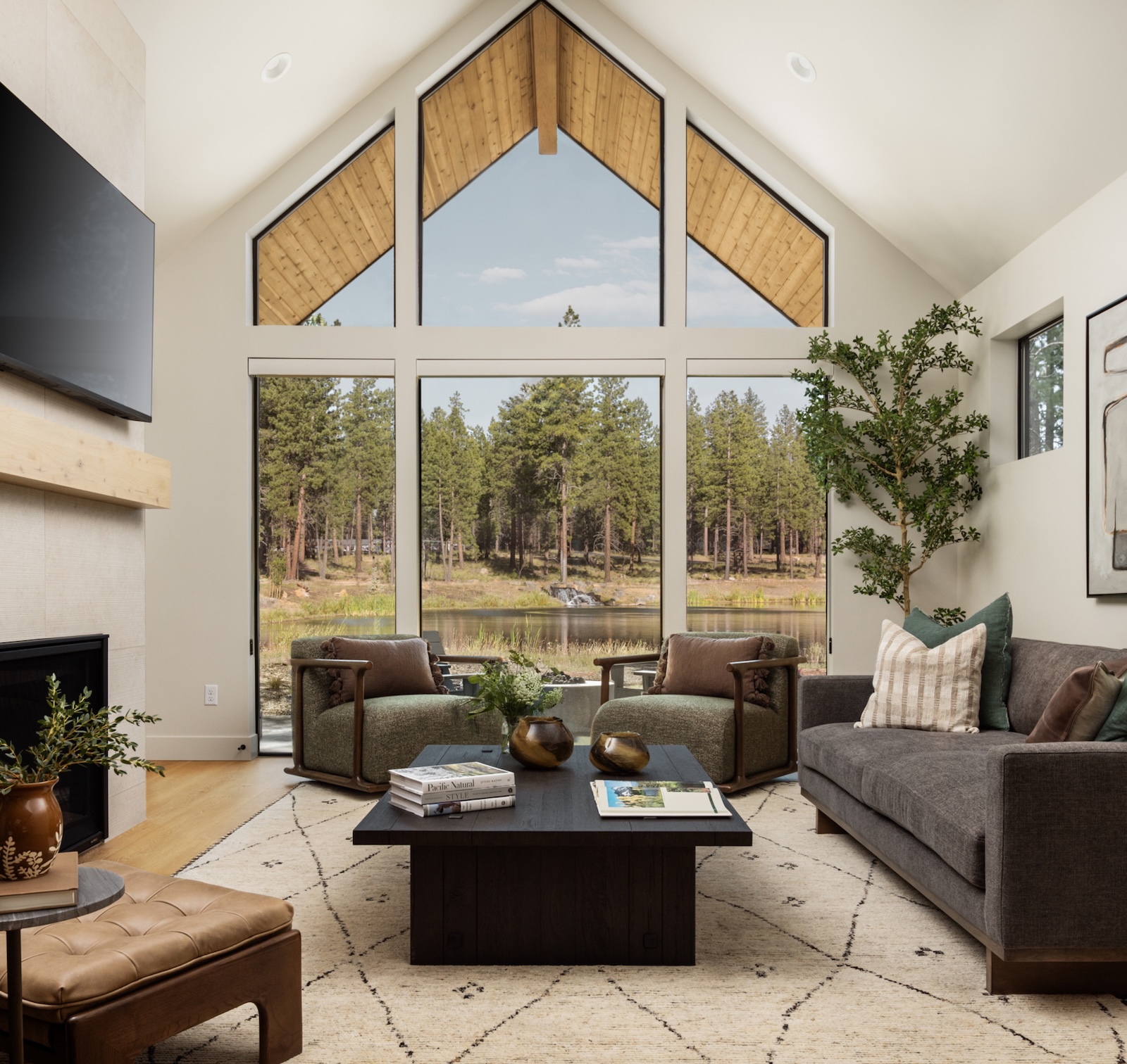 Vaulted living room with floor-to-ceiling windows overlooking a wooded pond, featuring a fireplace, neutral sofa, armchairs, and dark wood coffee table.